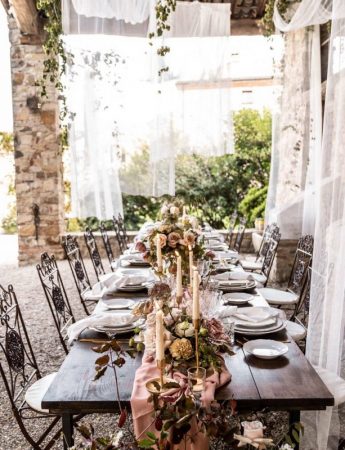 In questa foto un allestimento di nozze in stile Shabby Chic in campagna. Un runner decorato con fiori rosa e bianchi decora un tavolo di legno nudo. Su entrambi i lati sono disposti i piatti. Le sedie usate sono in ferro battuto