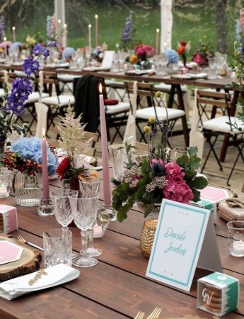 In questa foto una mise en place in stile Boho Chic. Piatti e posate sono disposti su tavoli imperiali in legno nudo senza tovagliato. Piccoli vasetti con fiori di campo sono usati come centrotavola abbinati a candele rosa