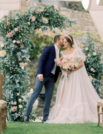In questa foto due sposi davanti ad un arco di fiori colore rosa e foglie. Lo sposo bacia sulla guancia la sposa che indossa un ampio
