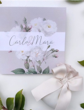 In questa foto un invito matrimonio realizzato con carta colore perla, fiori bianchi dipinti ad acquerello e, a chiudere, una striscia bianca di carta trasparente con i nomi degli sposi. Poggiato in basso a destra un pacchetto di confetti bianco