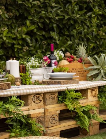In questa foto una mise en place realizzata su tavole di legno con piante e frutta, perfetta per un ricevimento green. I sottopiatti sono realizzati con tronchi e al centro del tavolo sono disposte candele bianche