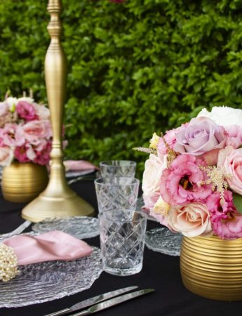 In questa foto una mise en place con tovaglioli rosa e centrotavola di rose in coordinato. I piatti sono trasparenti e i centrotavola con vasetti e alzatine colore oro